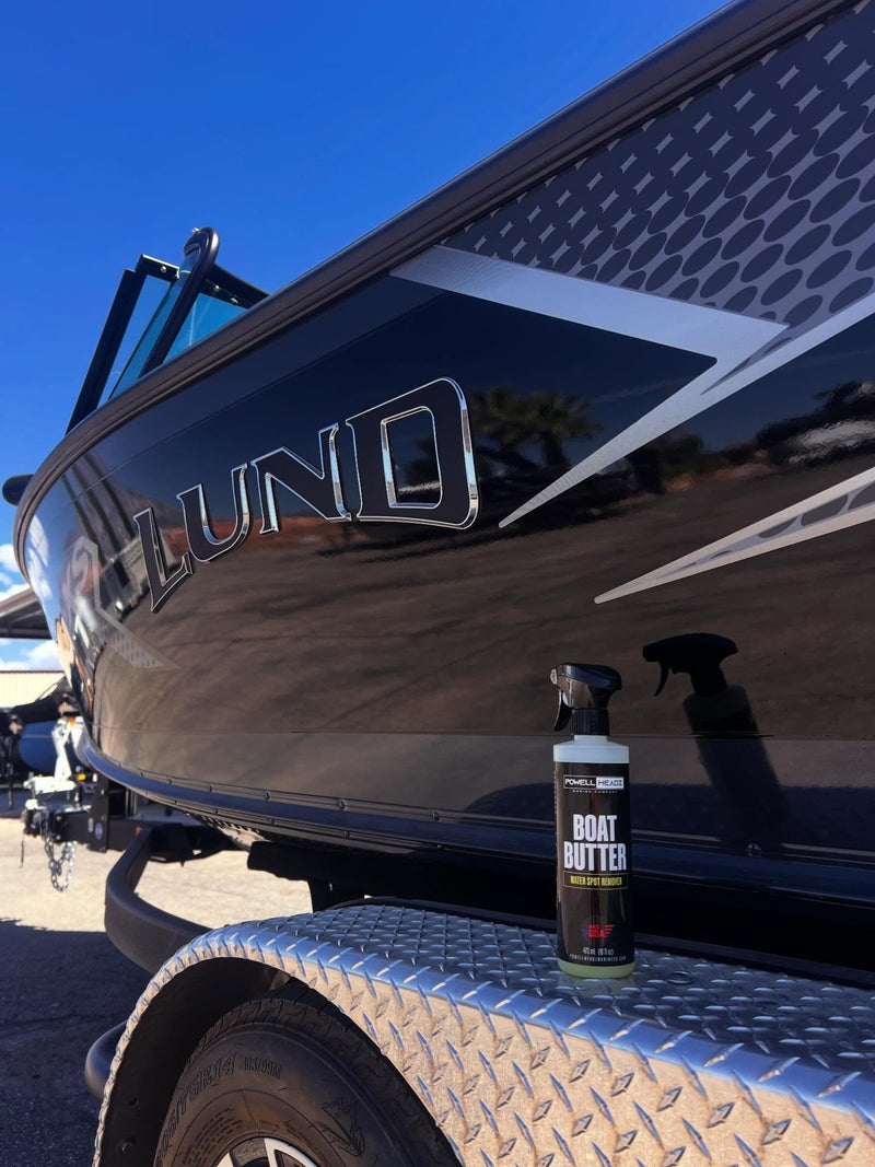 A genuine Boat Butter bottle stands on a trailer fender beside a dark Lund boat outdoors.
