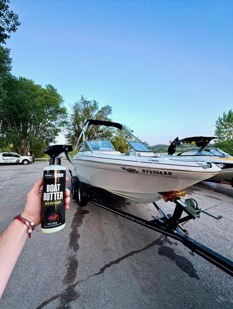 A person holding a Boat Butter spray bottle in front of a white boat on a trailer outdoors.